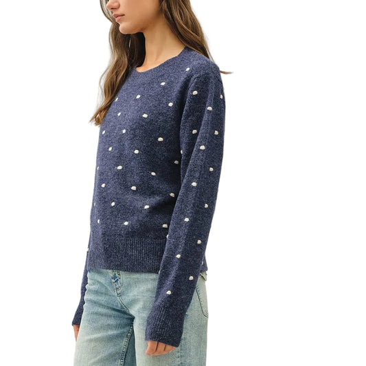 A woman modeling a navy blue knit sweater with small white embroidered dots, paired with light-wash wide-leg jeans, standing against a plain off-white studio background.