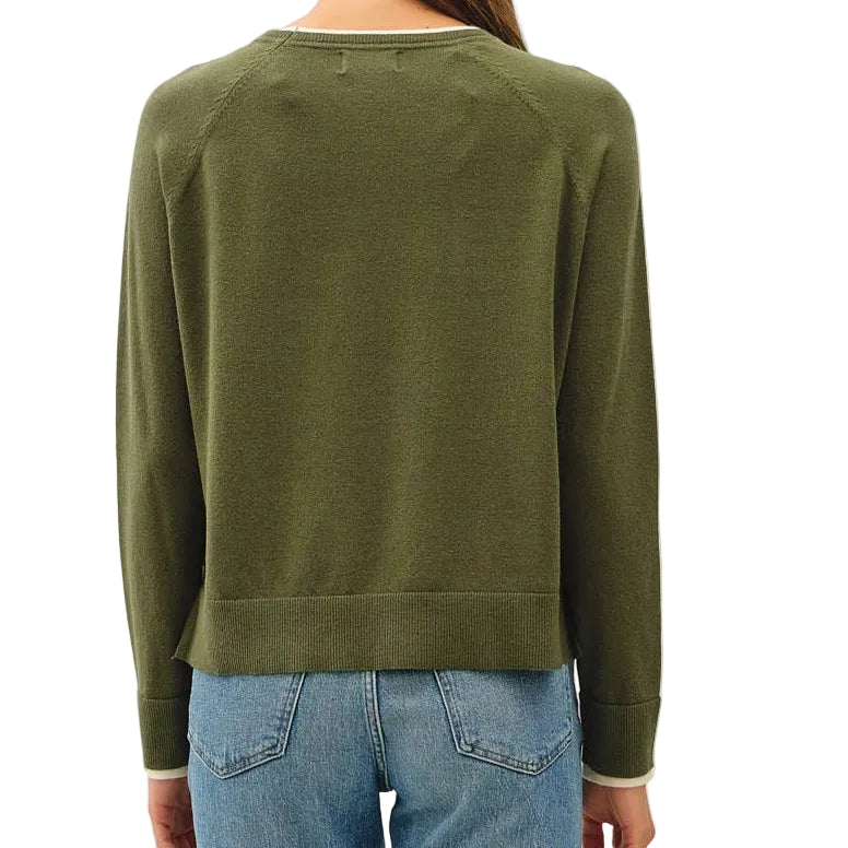 A woman viewed from behind wearing an olive green long-sleeve knit sweater, light blue cropped jeans, and tan slip-on shoes against a plain off-white studio background.