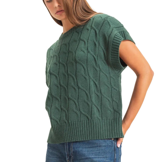 A woman wearing a dark green short-sleeve cable-knit sweater and blue jeans, standing against a plain off-white studio background.