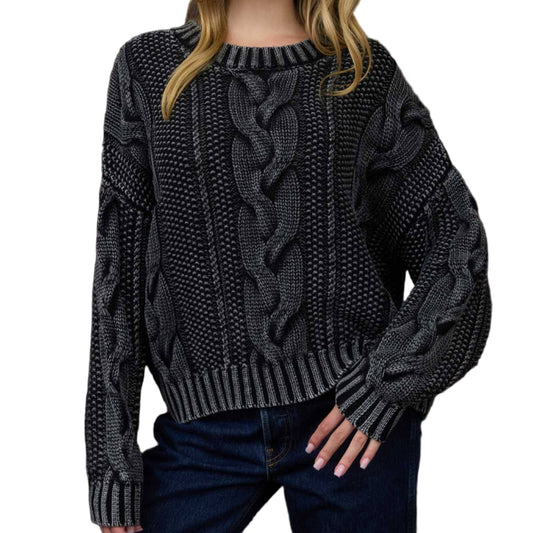 Woman wearing a dark gray cable knit sweater against a wooden paneled wall.