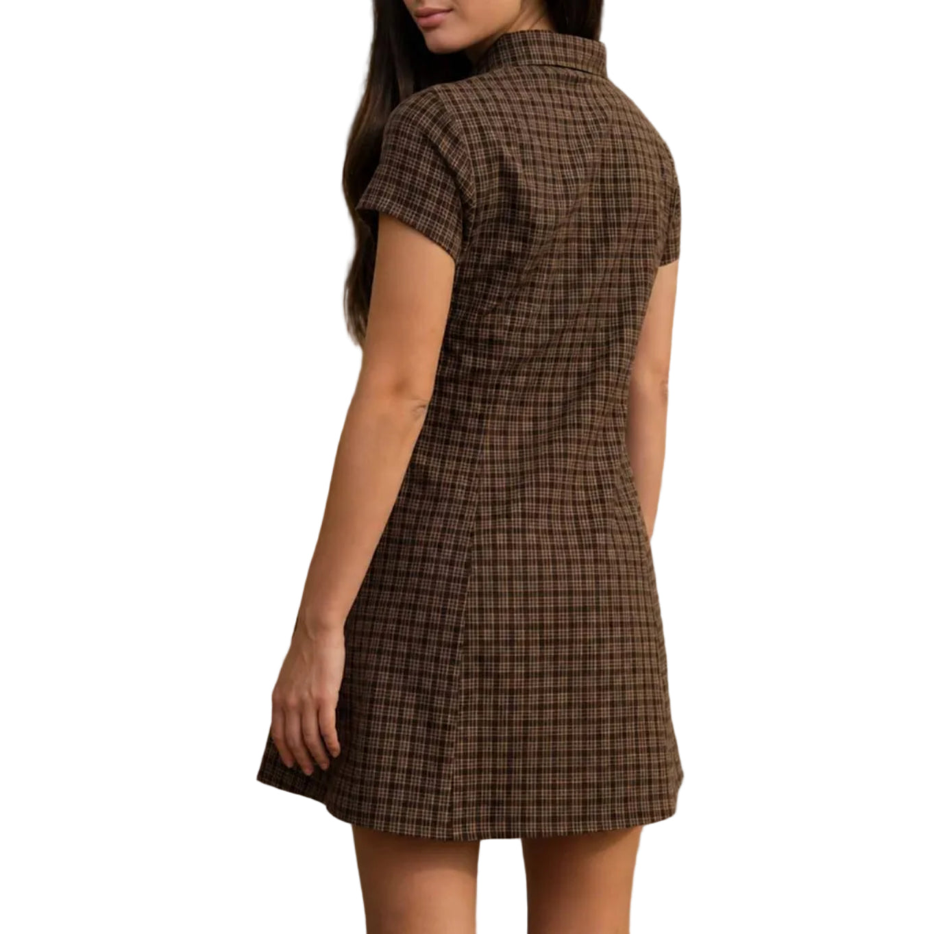 A woman wearing a brown and black checkered mini dress stands on a concrete path next to a large concrete wall, looking back over her shoulder in a desert landscape with mountains in the background.