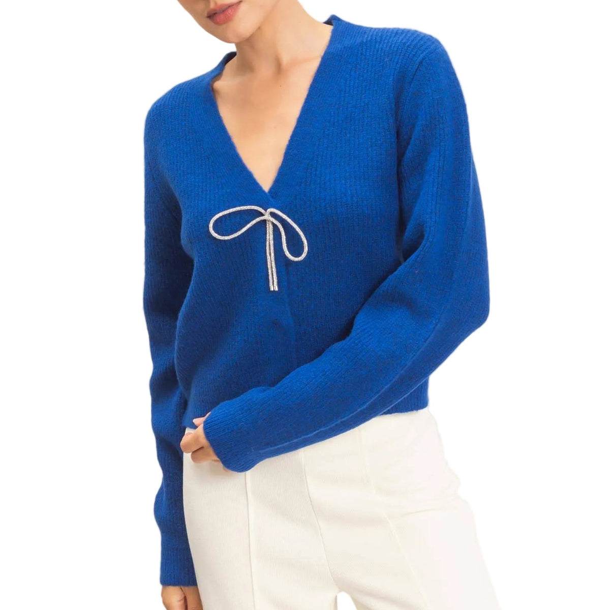 A woman wearing a bright royal blue ribbed knit sweater with a white embroidered bow detail on the chest, paired with white trousers, set against a plain light beige background.