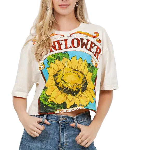 Woman wearing a graphic t-shirt with a sunflower design and blue jeans in a room with plants.