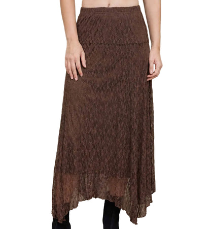 A woman wearing a brown lace midi skirt with a high-low hem, paired with black ankle boots, standing against a beige concrete wall.