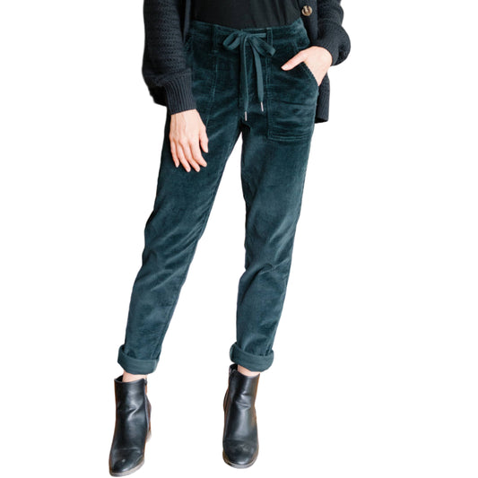 Woman in black top and green pants standing in a living room with a fireplace and dining table.