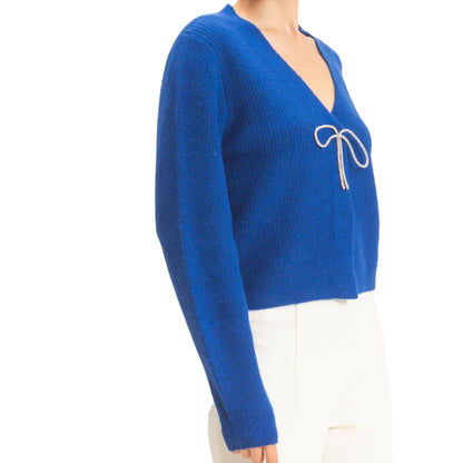 A woman modeling a royal blue ribbed knit sweater with a V-neck and a white bow detail on the chest, paired with white pants, against a plain off-white background.