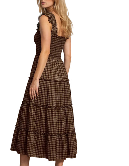A woman modeling a brown and black checkered tiered midi dress with ruffled shoulder straps, shown from a side-back angle against a plain white wall and gray floor.