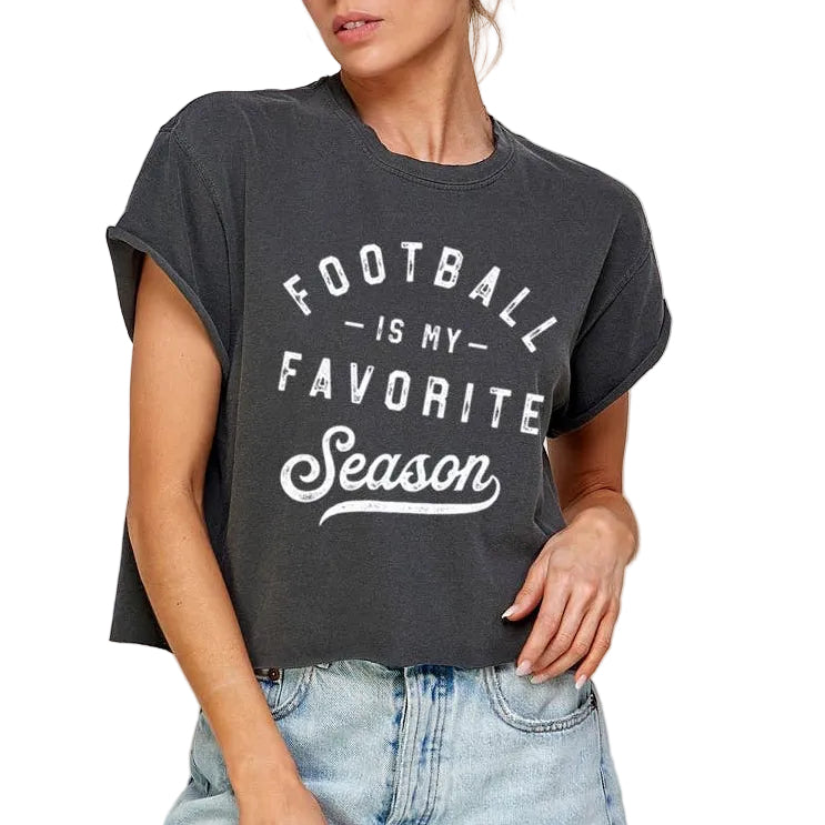 A woman wearing a dark charcoal gray graphic t-shirt with the text 'FOOTBALL IS MY FAVORITE Season' in white, paired with light-wash denim shorts, set against a plain light gray background.
