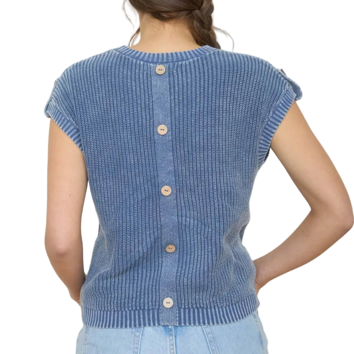 Woman wearing a blue ribbed knit sleeveless sweater with buttons, standing in front of framed artwork on a white wall.