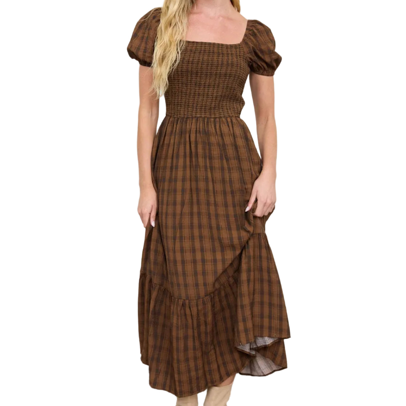 A woman modeling a brown and black plaid midi dress with puff sleeves, a smocked bodice, and a tiered ruffled hem, paired with beige ankle boots, standing against a white wall.