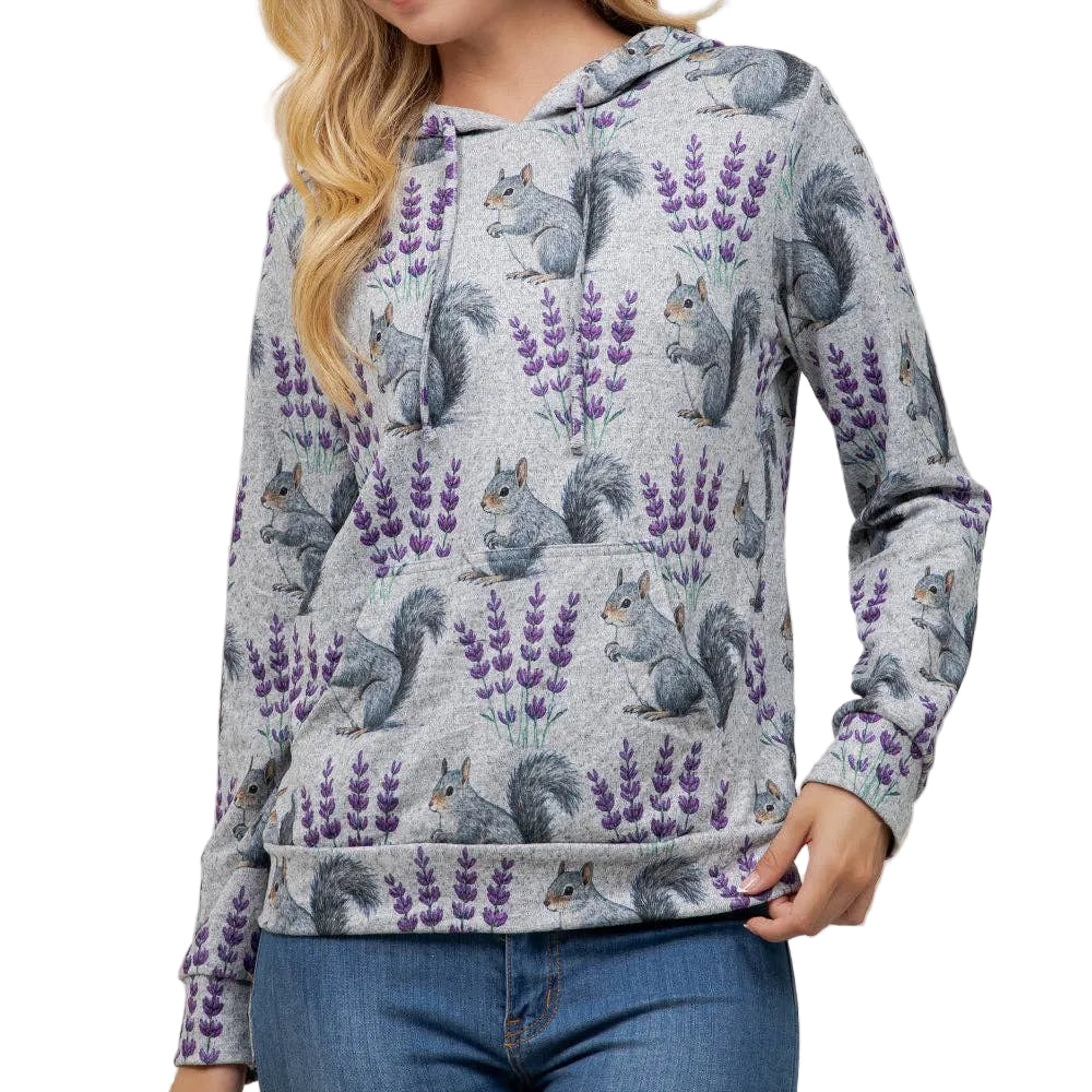 A woman wearing a light gray heathered hoodie with an all-over print of gray squirrels and purple lavender flowers, paired with blue jeans, standing against a plain off-white wall.