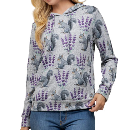 A woman wearing a light gray heathered hoodie with an all-over print of gray squirrels and purple lavender flowers, paired with blue jeans, standing against a plain off-white wall.