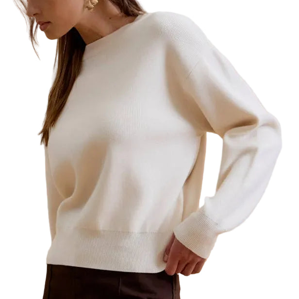 A woman modeling a white long-sleeve knit sweater and dark brown wide-leg trousers against a solid beige studio background.