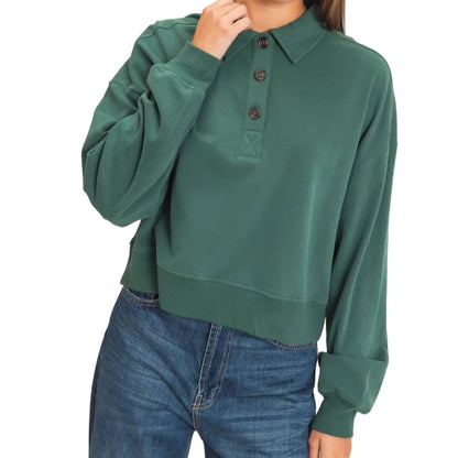 A woman wearing a dark green long-sleeve polo-style top with a button placket and blue jeans, standing against a plain light beige studio background.