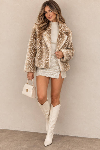 A woman models a beige faux fur jacket with white spots, styled with a white mini dress, white cowboy boots, and a small white handbag against a neutral beige wall.