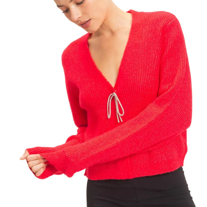 A woman wearing a bright red ribbed knit sweater with a deep V-neck and a small white drawstring detail at the center, paired with black pants, against a light beige studio background.