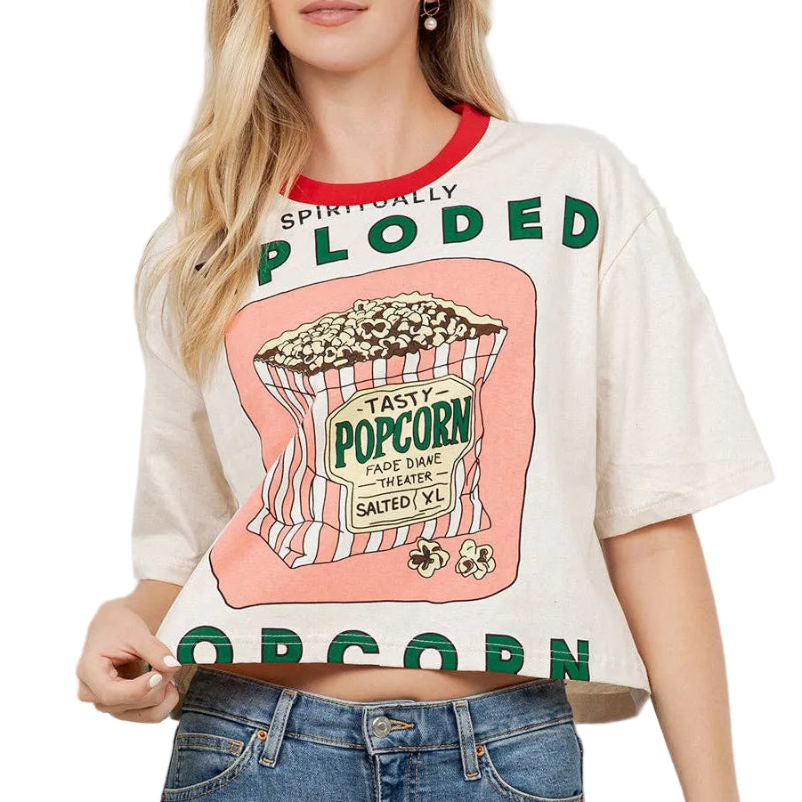 Woman wearing a graphic t-shirt with 'Plooded Popcorn' design and blue jeans, standing against a white wall.