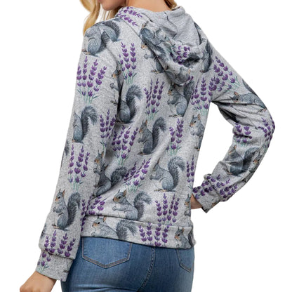 A woman wearing a light gray hooded sweatshirt with an all-over print of purple lavender and gray squirrels, paired with blue jeans, shown from a side-back angle against a plain off-white wall.