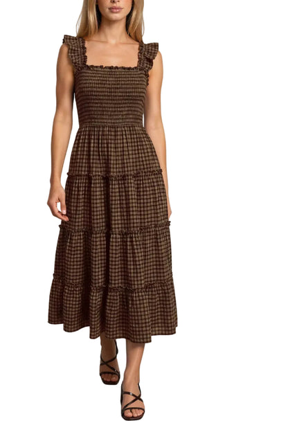 A woman modeling a brown and black checkered tiered midi dress with a smocked bodice and ruffled cap sleeves, paired with brown strappy sandals, against a neutral studio backdrop.