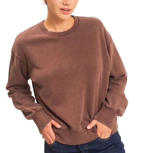 A woman modeling a brown long-sleeve crewneck sweatshirt with a faded wash, paired with a dark denim mini skirt, against a plain light beige background.