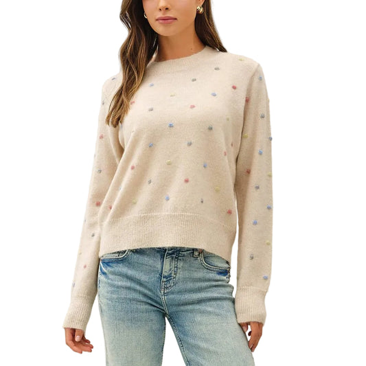 A woman modeling a cream-colored knit sweater with small multicolored embroidered dots, paired with light-wash flare jeans and brown shoes, against a plain white studio background.