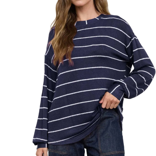 A woman wearing a navy blue and white horizontally striped knit sweater with dark blue jeans, standing against a light gray studio background.