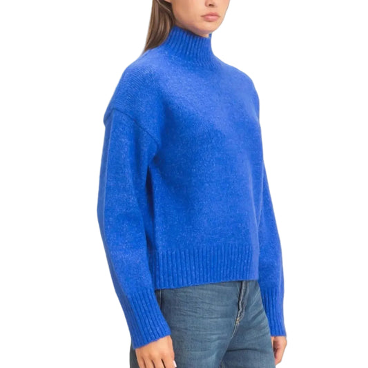 A woman wearing a bright royal blue turtleneck sweater and blue jeans, standing against a plain off-white studio background.
