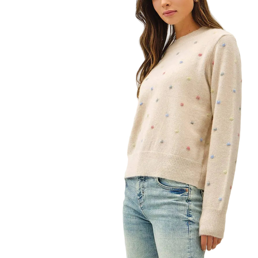 A woman modeling a cream-colored knit sweater with small colorful embroidered details, paired with light-wash blue flare jeans and tan shoes, against a plain white studio background.