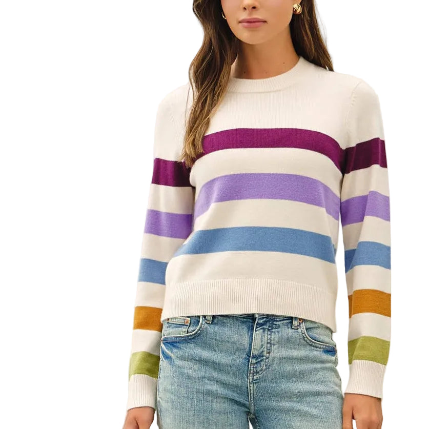 A woman modeling a cream knit sweater with horizontal purple, blue, and orange stripes on the sleeves, paired with light-wash flare jeans and tan shoes, against a plain white studio background.