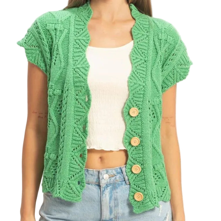 Woman wearing a green knitted cardigan over a white top and denim shorts on a plain background