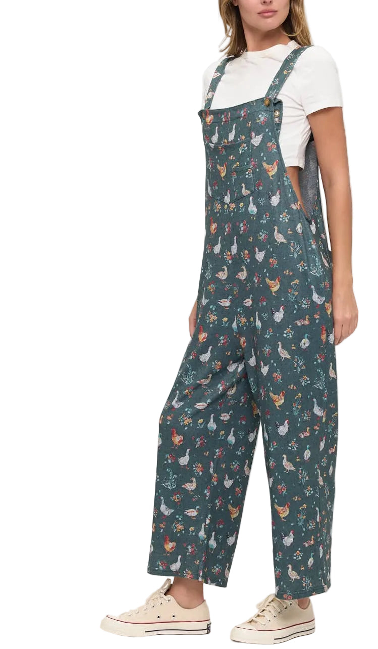 A woman modeling dark teal wide-leg overalls with a colorful woodland animal print, styled with a white t-shirt and beige sneakers against a plain white studio background.