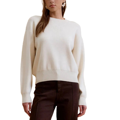 A woman modeling a white crewneck knit sweater and dark brown wide-leg trousers against a solid beige studio background.