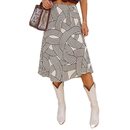 A woman stands outdoors wearing a maroon graphic T-shirt with 'TEXAS' in white, a black-and-white geometric print midi skirt, and white cowboy boots. She is holding a clear handbag and posing against a beige wall with a sidewalk and storefront signs in the background.