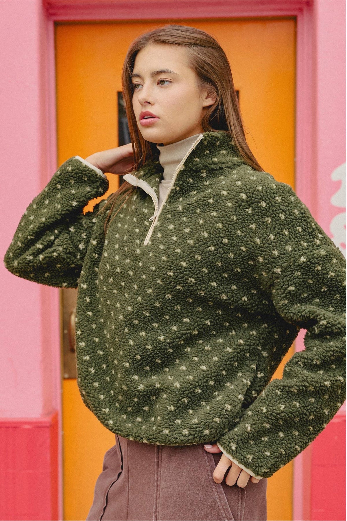 A woman wearing a dark olive green fleece quarter-zip pullover with a small beige floral pattern, posed in front of a vibrant pink and orange doorway.