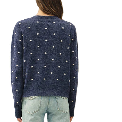 A woman viewed from behind wearing a navy blue knit sweater with white polka dots and light-wash wide-leg jeans, standing against a plain off-white wall.