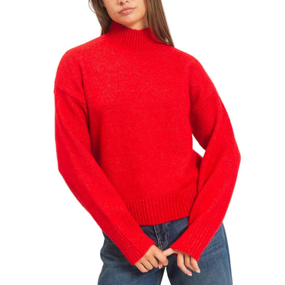 A woman wearing a bright red knit mock neck sweater and blue jeans, standing against a plain light beige studio background.