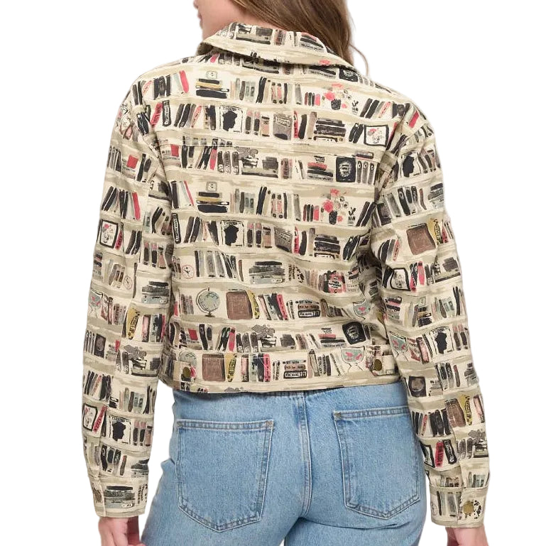 A woman viewed from behind wearing a cropped jacket with an all-over bookshelf print, paired with light blue jeans, standing in a brightly lit room next to a vase with dried branches.