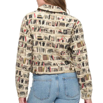A woman viewed from behind wearing a cropped jacket with an all-over bookshelf print, paired with light blue jeans, standing in a brightly lit room next to a vase with dried branches.