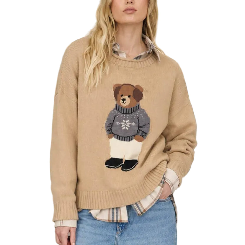 A woman stands on a city street wearing a beige crewneck sweater with a teddy bear graphic, blue jeans, and brown sneakers, while holding a leash with a small brown dog.