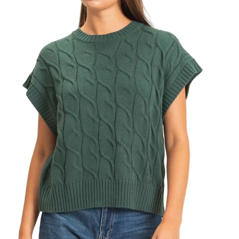 A woman modeling a dark green short-sleeve cable-knit sweater with blue jeans and beige shoes, standing against a plain light beige studio background.