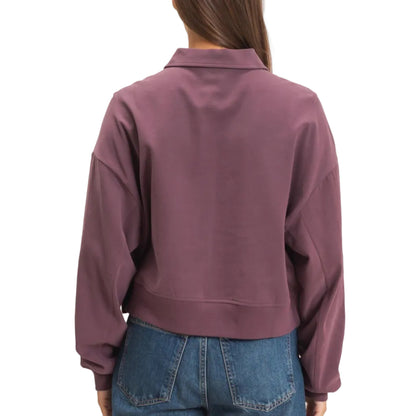 A woman viewed from the back wearing a cropped, long-sleeve collared sweatshirt in a muted mauve-purple color, paired with blue jeans, against a plain light beige background.