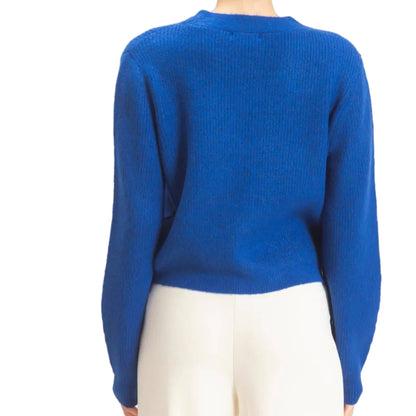 A woman viewed from the back wearing a bright royal blue ribbed knit sweater and white pants against a plain light background.