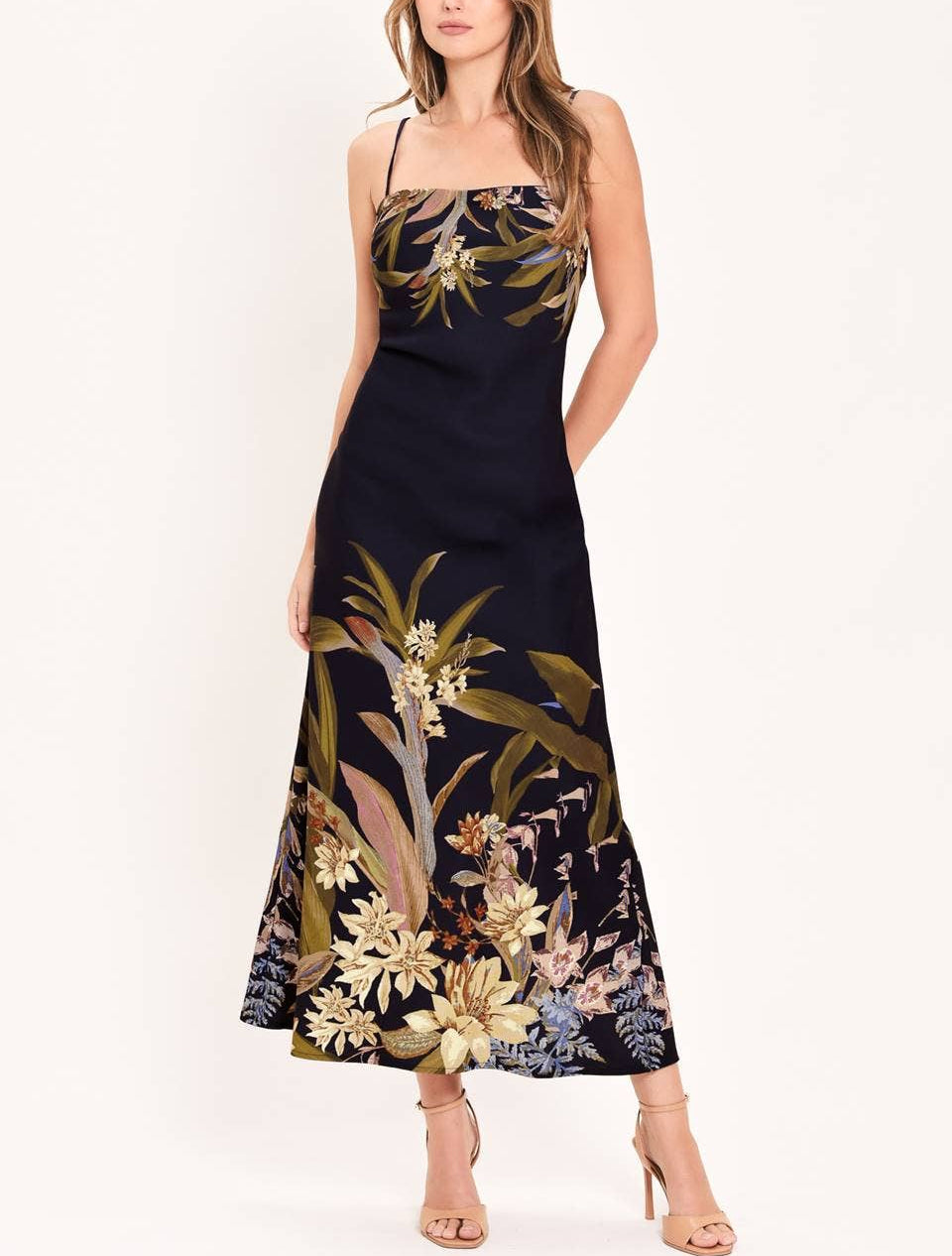 A woman modeling a sleeveless navy maxi dress with a tropical floral and botanical print in beige, gold, and muted green, paired with nude high-heeled sandals against a plain white studio background.