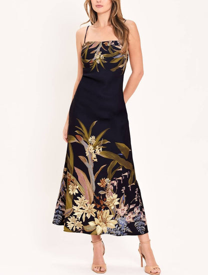 A woman modeling a sleeveless navy maxi dress with a tropical floral and botanical print in beige, gold, and muted green, paired with nude high-heeled sandals against a plain white studio background.