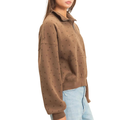 A woman wearing a brown cropped quarter-zip sweatshirt with a subtle polka dot pattern, paired with light blue jeans, photographed against a plain off-white studio background.