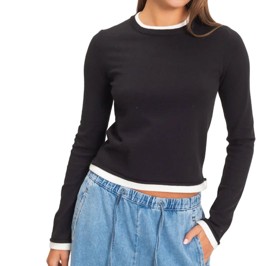 A woman modeling a black long-sleeve crewneck sweater with white ribbed trim at the collar, cuffs, and hem, paired with light blue jeans against a plain light beige background.