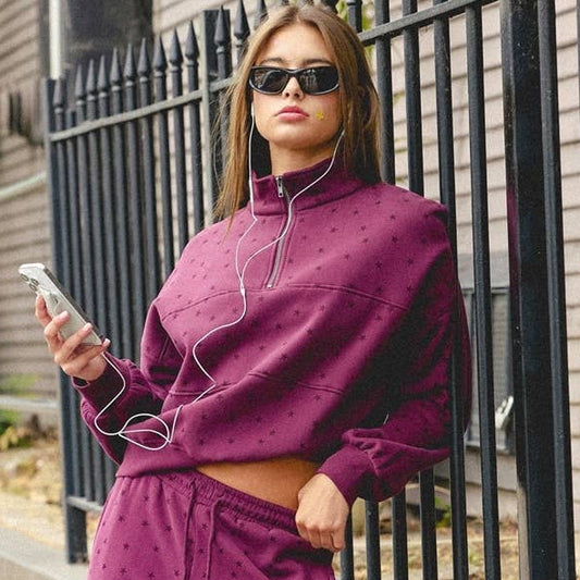 A woman stands on a sidewalk wearing a matching two-piece lounge set in a deep berry purple. The set consists of a cropped long-sleeve top and wide-leg pants, both featuring a subtle dotted pattern.