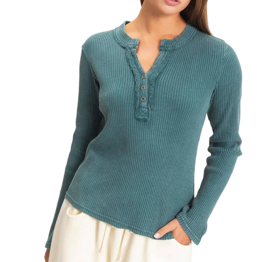 A woman wearing a teal ribbed long-sleeve henley top with a button placket, paired with cream pants, standing against a plain light beige background.
