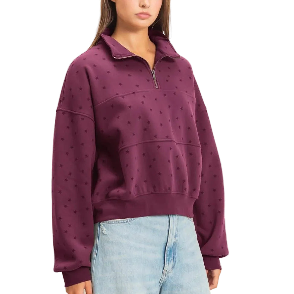 A woman modeling a deep purple half-zip sweatshirt with a subtle dotted pattern, paired with light-wash wide-leg jeans and brown suede boots, against a plain beige studio background.