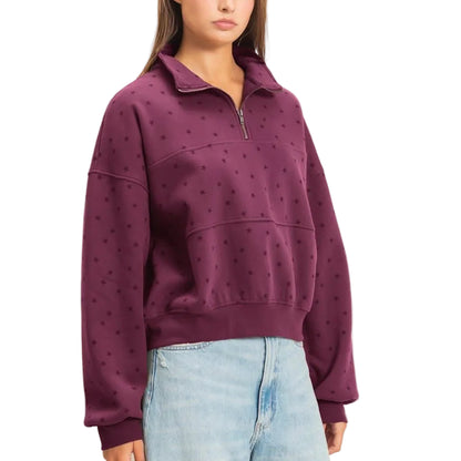 A woman modeling a deep purple half-zip sweatshirt with a subtle dotted pattern, paired with light-wash wide-leg jeans and brown suede boots, against a plain beige studio background.